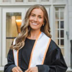Female UT student wearing graduation attire standing and smiling in front of an academic building.