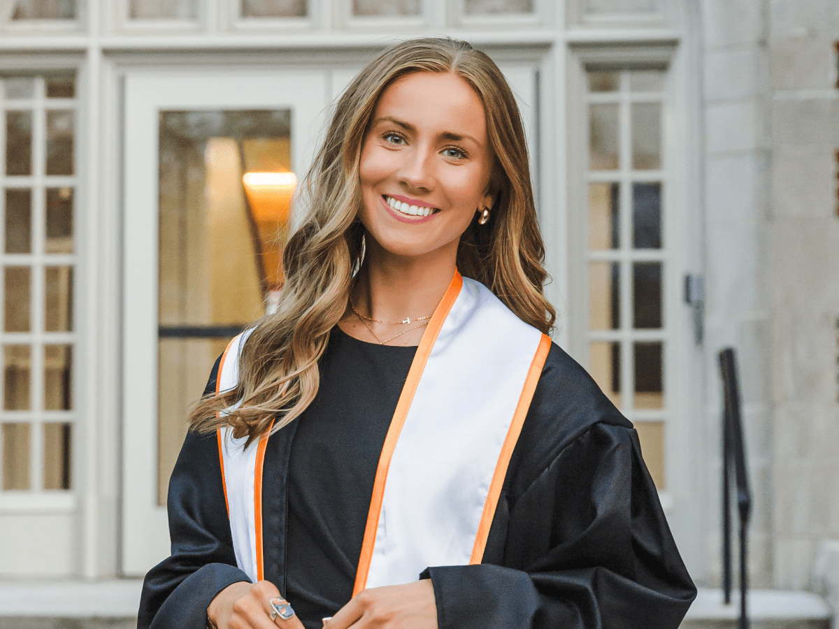 Female UT student wearing graduation attire standing and smiling in front of an academic building.