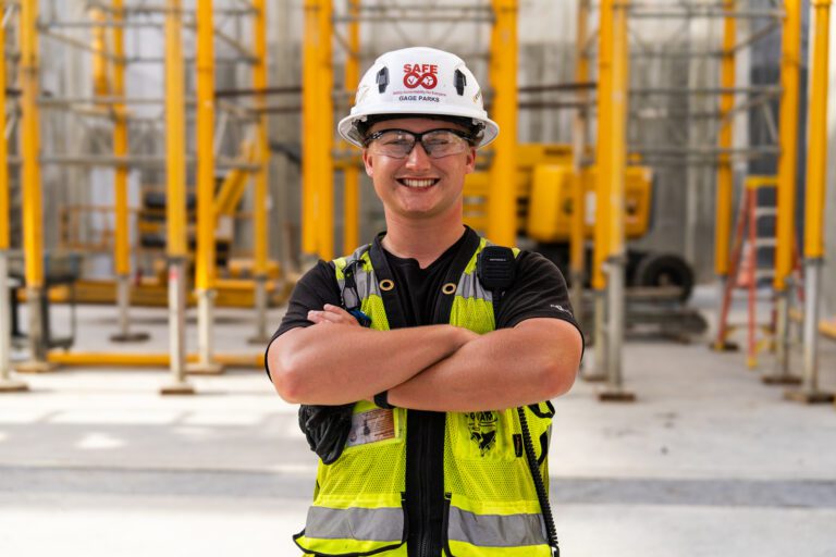 Gage Parks smiling with arms crossed wearing a hard helmet and yellow vest in front of a construction project