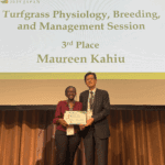 Female PhD student being presented with an award on a stage with the words "Turfgrass Physiology, Breeding, and Management Session 3rd Place Maureen Kahiu"