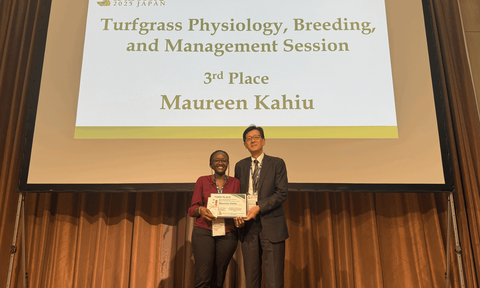 Female PhD student being presented with an award on a stage with the words "Turfgrass Physiology, Breeding, and Management Session 3rd Place Maureen Kahiu"