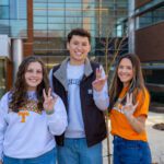 Three students wearing Tennessee attired, smiling and holding up the "Vols" sign outside am academic building.