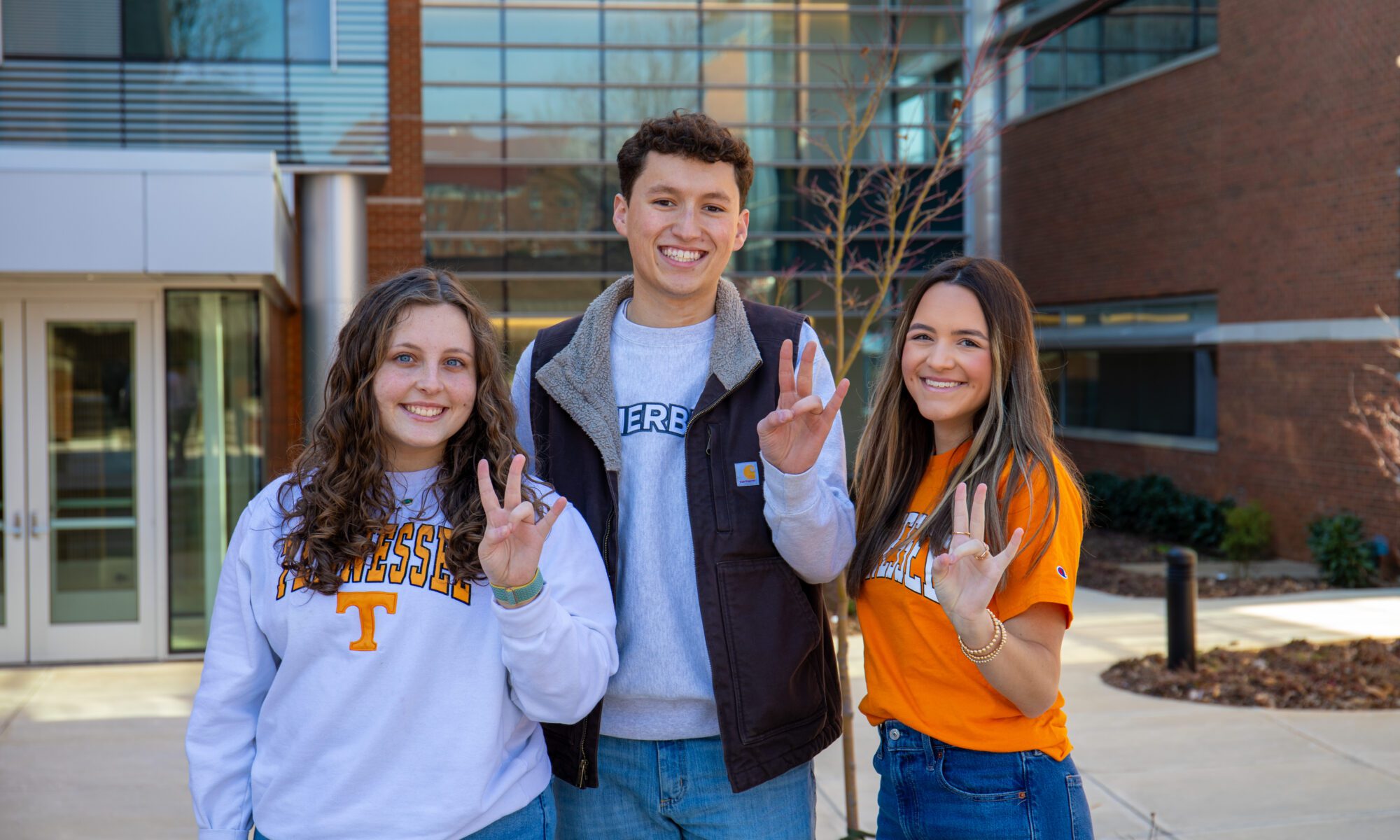 Three students wearing Tennessee attired, smiling and holding up the "Vols" sign outside am academic building.
