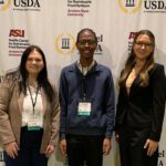 Three students dressed in business professional attire standing in front of a Model USDA backdrop.