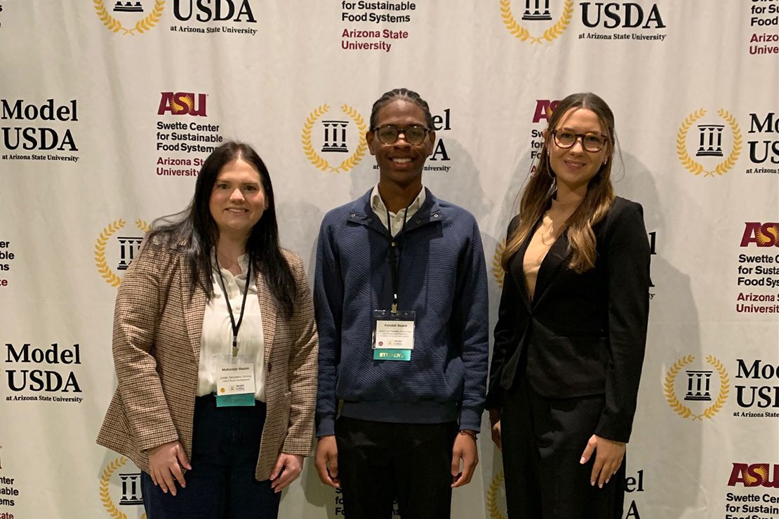 Three students dressed in business professional attire standing in front of a Model USDA backdrop.